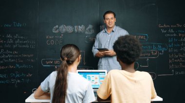 Kafkasyalı öğretmen elinde tablet tutarken öğrenci sorusuna cevap veriyor. Beyaz kız, BTMM teknoloji sınıfında dizüstü bilgisayar ekranı görüntülerken kod ile tahtaya işaret ediyor. Öğretim.