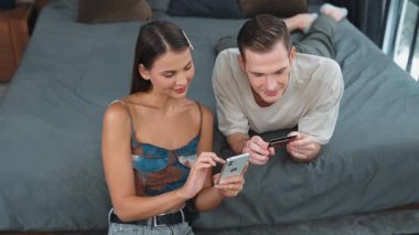Genç çift, kredi kartıyla ödemek için internet üzerinden ödeme uygulaması ve dijital cüzdan kullanarak yatak odasında oturuyor. Mobil internet üzerinden alışveriş ve modern alımlar. Düzenle