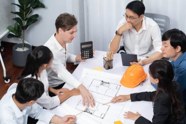 Mimari proje üzerinde birlikte çalışan çeşitli inşaat mühendisi ve müvekkil grupları, inşaat planını gözden geçirip toplantı masasında plan oluşturuyorlar. Tedbirli