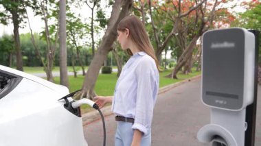 Yavaş çekimde genç bir kadın sonbaharda ulusal parktaki EV şarj istasyonunda EV elektrikli arabaları şarj ediyor. Tatil ve sonbahar sezonunda çevre dostu arabalarla seyahat. Yararlı