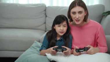 Mutlu anne ve enerjik kız oyun oynuyor ve birlikte vakit geçiriyorlar. Kafkasyalı anne, elinde joystick tutarken çocuğunun yanında oturuyor ve rahat oturma odasında kızıyla oyun oynamaya odaklanıyor. Pedagoji.