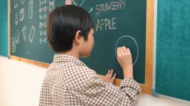 Asyalı çocuk sınıfta tahtada dururken hayal gücünü ifade etmek ve geliştirmek için resim çiziyor. Sanat dersinde karatahtaya yazı yazan çekici ilkokul öğrencisi. Eğitim konsepti. Pedagoji.