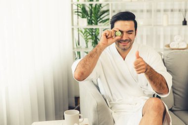 Elinde taze salatalık dilimleri tutan ve bornoz giyen bir adam tatil köyünde ya da otelde yüz bakımının keyfini çıkarıyor. Yüz ve güzellik tedavisi için cilt bakımı. Sessiz.