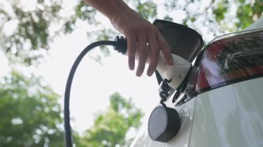 Yavaş çekim adamı, yaz mevsiminde açık yeşil yay yeşilliğindeki şarj istasyonundan elektrikli araba aküsünü şarj etmek için EV şarj aleti taktı. Yeniden şarj edilebilir elektrikli araç için sürdürülebilir enerji