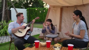 Baba gitar çalarken anne ve kız birlikte kampta oturuyorlar. Ebeveynler, genç nesil çapraz kuşak farklılıklarıyla zaman geçirmek için açık hava kamp aktivitelerini kullanıyorlar. Uyumsuzluk.