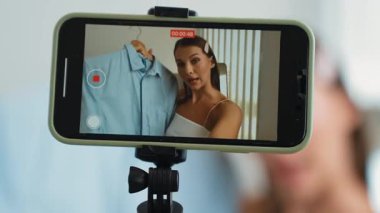 Genç sosyal medyum kadın moda videosu yapıyor. Blogcu kameraya selfie çubuğu ve ışık halkası kullanarak gülümserken, aynı zamanda ikna edici çevrimiçi kıyafetlerin izleyicilere veya takipçilere vlog satmasını sağlıyor. Düzenle