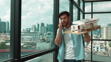 Gülümseyen Kafkasyalı mühendis ev modelini sağa sola çeviriyor ve iş arkadaşına telefon ederken hata noktasını kontrol ediyor. Profesyonel yönetici teftiş, kontrol, mimari modele bakmak. İzleme