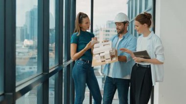 Proje yöneticisi ev modelini elinde tutarken, mimar mühendis takımı da ev tasarımı veya bina inşaatı hakkında fikir alışverişinde bulunuyor. Şehrin penceresinin yanında iç mimar duruyor. İzleme