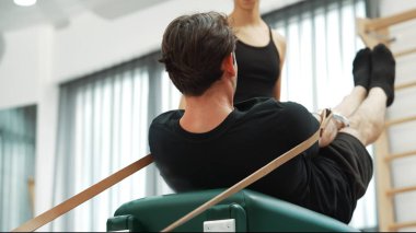 Müşteri, eğitmen desteğinin rehberliğinde kayışlarla Pilates reformatında egzersiz yapıyor. Sağlıklı bir adam pilates yapıyor ve üst vücut geliştirmek için Cadillac 'a asılıyor. Alışkanlık.