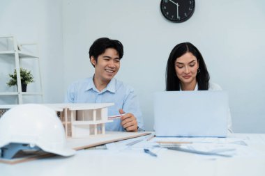 Profesyonel Asyalı erkek mimar ev modelinin uzunluğunu ölçmek için cetvel kullanırken, beyaz tenli genç meslektaşı ev modeliyle toplantı masasındaki verileri analiz etmek için dizüstü bilgisayar kullanıyor. Tertemiz..