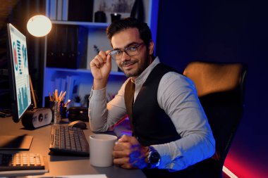 Masada uzaktan çalışan gözlüklü akıllı iş adamı profili, yelek kostümlü kameraya bakmak, neon karanlık aydınlatma ofisinde profesyonel pazarlama sunumu. Tahmin et..