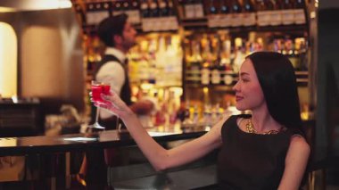 Güzel bir konuk, gece kulübünün mükemmel atmosferinde barmenlerin arka planında çiçeklerle süslenmiş karışık votka Cosmopolitan kırmızı kokteyli ile lüks bir gişede içki içerken kameraya bakıyor. Vinosity.