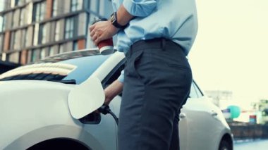 Takım elbiseli, hırslı bir işadamı elektrik kablosu ve yenilenebilir enerji enerjili bir elektrikli araçla şarj istasyonunda dururken elektrikli aracına yaslanıyor..