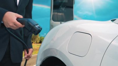 İlerici işadamı elektrikli arabayı tepe üzerindeki yolculuğunda şarj istasyonuyla dolduruyor. Temiz çevre için yenilenebilir enerji ile çalışan EV arabası. EV otomobilinden enerji sürdürülebilirliği kavramı.