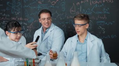 Laboratuvar önlüğü giyen genç bir çocuk, STEM fen sınıfında tahtada renkli sıvıyla bekletiliyor. Öğretmen masada tavsiye verirken bir grup farklı öğrenci deney yapıyor. Öğretim