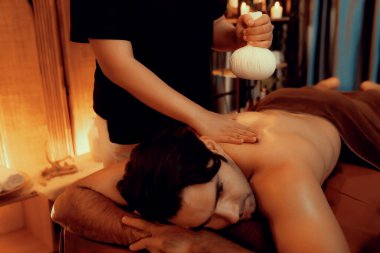 Sıcak bitkisel top spa masaj vücut tedavisi, masör erkek vücuduna nazikçe şifalı bitki torbası bastırıyor. Aromaterapi tesisinin sakinliği ve dinginliği kaplıca salonundaki mumların sıcacık ışığında. Sessiz.