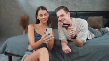 Genç çift, kredi kartıyla ödemek için internet üzerinden ödeme uygulaması ve dijital cüzdan kullanarak yatak odasında oturuyor. Mobil internet üzerinden alışveriş ve modern alımlar. Düzenle