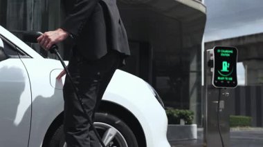 İşadamı EV şarj aletini şehir merkezindeki EV şarj istasyonundan ya da otoparktan şarj etmek için takıyor. Çevre dostu şarj edilebilir araba şehir yaşamında alternatif enerji kullanıyor. İnceleyin.