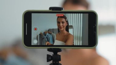 Genç sosyal medyum kadın moda videosu yapıyor. Blogcu kameraya selfie çubuğu ve ışık halkası kullanarak gülümserken, aynı zamanda ikna edici çevrimiçi kıyafetlerin izleyicilere veya takipçilere vlog satmasını sağlıyor. Düzenle