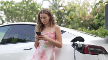 Genç bir kadın doğadaki EV şarj istasyonunda elektrik için akıllı telefon kullanıyor. Modern çevre ve EV araçlı sürdürülebilir otomobil taşıma yaşam tarzı. Titiz