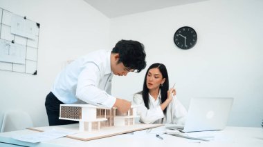 Profesyonel Asyalı erkek mimar ev modelinin uzunluğunu ölçmek için cetvel kullanırken, beyaz tenli genç meslektaşı ev modeliyle toplantı masasındaki verileri analiz etmek için dizüstü bilgisayar kullanıyor. Tertemiz..