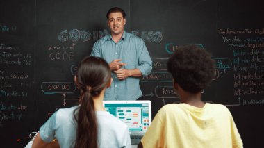 Tahtada çeşitli öğrencilere mühendislik kodları öğreten yakışıklı bir öğretmen. Mutlu çok kültürlü liseli çocuklar dizüstü bilgisayar kullanırken öğreniyor ya da programlama veya kodlama sistemini öğreniyor. Öğretim.
