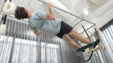 Çekirdek dayanıklılığı ve spor salonunda vücut egzersizi için pilates trapez masasına tırmanan bir adam. Eğitmen, arka ve kalça esnekliğini arttırmak için Cadillac 'ta ters asılı duruyor. Alışkanlık.