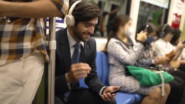 Gülümseyen yakışıklı yatırımcı trende otururken rahatlatıcı müzik dinliyor. Profesyonel iş adamı iş yerine toplu taşıma araçlarıyla giderken müzik dinlemeyi sever. Arka plan bulanık. Sevinçli..