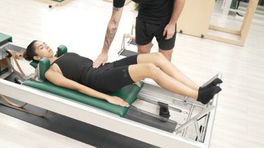 Mutlu pilates eğitmeni, bir müşteriye çekirdek istikrarı geliştirmek için bir reformcu üzerinde bacak egzersizi yapmasında yardımcı oluyor. Pilates makinesi kullanan sportif kız ark fıçısında kol dairesi pozu veriyor. Alışkanlık.