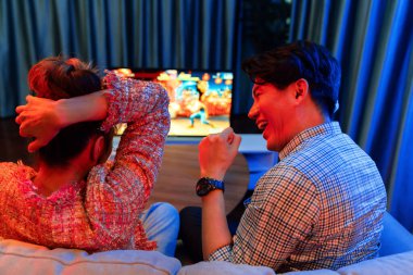 joystickli bir çift, TV ekranında dövüş oyunu oynuyor, erkek, kız arkadaşıyla yarışma seviyesini geri kazanıyor, modern evdeki neon ışıklı oturma odasında kanepeye oturuyor. İnfobahn.