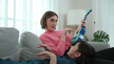 Kanepede annesinin kucağında yatarken Asyalı tatlı kız akustik müzik dinliyor. Beyaz bir anne renkli ukulele çalarken tatlı bir çocuk ailesine bakıyor. Aile eğlence konsepti. Pedagoji.