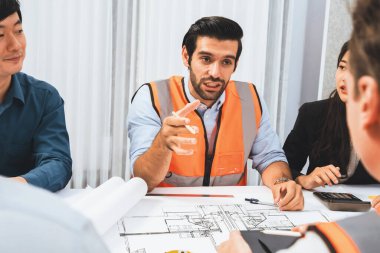 Mimari proje üzerinde birlikte çalışan çeşitli inşaat mühendisi ve müvekkil grupları, inşaat planını gözden geçirip toplantı masasında plan oluşturuyorlar. Tedbirli