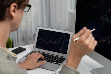 Stresli iş adamı günü modern ofisteki dinamik borsa grafiğine bakıyor, veri tabanı bilgisayarını ve bilgisayarını evden çalışırken karşılaştırıyor. Bireysel.