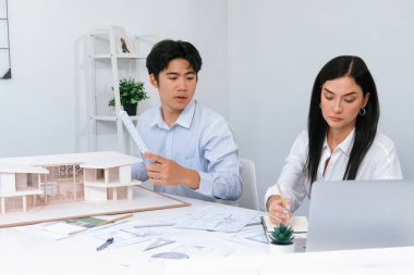 Profesyonel Asyalı erkek mimar ev modelinin uzunluğunu ölçmek için cetvel kullanırken, beyaz tenli genç meslektaşı ev modeliyle toplantı masasındaki verileri analiz etmek için dizüstü bilgisayar kullanıyor. Tertemiz..