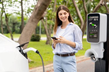 Ulusal Park veya Yaz Ormanı 'ndaki tatil gezisi sırasında EV şarj istasyonundan elektrik aküsü almak için akıllı telefon internet bankacılık uygulaması kullanan genç bir kadın. Yükselt