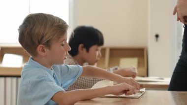 Beyaz tenli ilkokul öğrencisinin tahtada otururken bir nota bakması ve soruya cevap vermesi. Çocuklar derse katılırken genç öğretmen sınıf çalışmasını ya da sınavı açıklar. Pedagoji.