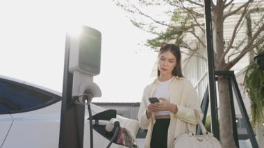 Ağır çekimde genç bir kadın, alışveriş merkezinin otoparkındaki şarj istasyonundan şarj olan EV 'nin pilini şarj ederken akıllı telefondan akü durumunu kontrol ediyor. Modern kadın elektrikli araçla alışverişe gider. Yararlı