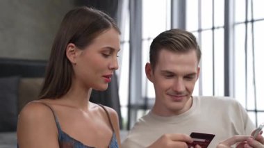 Genç çift, kredi kartıyla ödemek için internet üzerinden ödeme uygulaması ve dijital cüzdan kullanarak yatak odasında oturuyor. Mobil internet üzerinden alışveriş ve modern alımlar. Düzenle