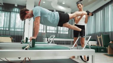 Yetenekli pilates eğitmeni, yenilikçi bir makinede bacak uzantısı olan bir tahtada müşteriye yardımcı olur. Bayan antrenör dengeyi geliştirmek için kalas değişimine yardımcı oluyor. Alışkanlık.