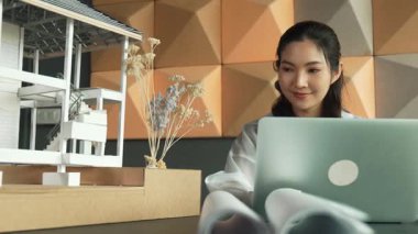 Genç ve güzel mimar mühendisin çatı malzemelerini kontrol etmesi ve ofisteki masaya yerleştirilmiş proje planı ve ev modeliyle dizüstü bilgisayarı kullanması. Yaratıcı iş tasarımı konsepti. Yönlendirici.