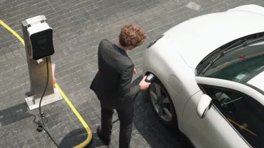 İlerici işadamı, şehir merkezinde dolaşmadan önce şarj cihazından elektrikli arabasına şarj prizi takıyor. Çevre dostu şarj edilebilir araba, sürdürülebilir ve temiz enerjiyle çalışıyor..