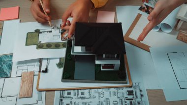 Bir evin detaylı bir mimari modelini gösteren yaratıcı bir çalışma alanı. Planlar ve tasarım materyalleri, işbirliği ve yenilikler üzerine tartışmalar yapan eller. SACTR