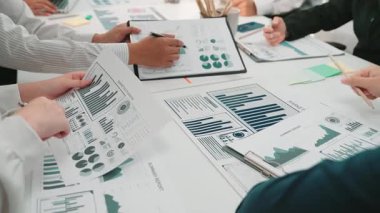 Çeşitli iş adamları, modern bir ofiste tablolar ve grafiklerle finansal verileri analiz ederek, takım çalışmalarını ve yenilikleri teşvik ederek, işbirlikçi bir toplantı düzenliyor. SACTR