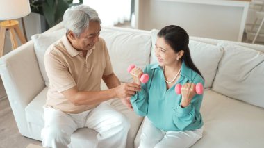 Mutlu yaşlı çift evde dambıllarıyla egzersiz yapıyor, kas gücü, eklem sağlığı ve sağlıklı yaşlanma için fiziksel sağlığa odaklanıyor. Emeklilik konsepti. Myrmidon.