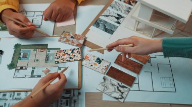 İç dizayn işleriyle uğraşan, tasarım planları ve örneklerle dolu bir masada materyal ve planların analiz edildiği dinamik bir sahne. SACTR