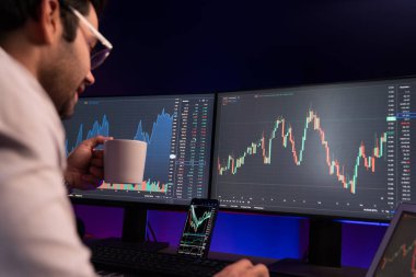 Zeki tüccar işadamı kahve fincanı içiyor, neon ışıklı ofisteki arama pazarında yüksek karlı finans teknolojisiyle bilgisayar üzerinde iki dinamik borsa yatırım ekranını analiz ediyor. Tahmin et..
