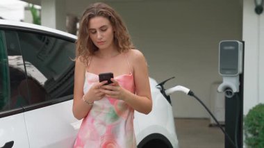 Modern çevre dostu kadın EV şarj istasyonundan elektrikli aracı şarj ediyor. Evde şarj edilen pilleri optimize etmek için enerji kullanımını takip etmek için yenilikçi EV teknolojisi kullanımı. Titiz