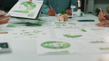 Bir iş ekibi parlak bir ofiste sürdürülebilirlik projesinde işbirliği yapıyor. Çeşitli grafikleri analiz edip yenilikçi fikirleri paylaşırken, çevre dostu kavramları vurguluyorlar. SACTR