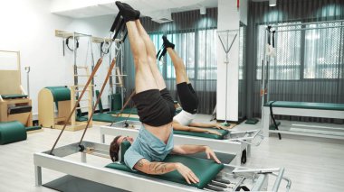 Müşteri, spor salonundaki yenilikçi bir makinede kayışla ters bacak kaldırma hareketi yapıyor. Çekirdek bağlantısını, dengesini ve esnekliğini geliştirmek için ters takla atan iki mutlu antrenör. Alışkanlık.