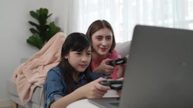 Annem dizüstü bilgisayar kullanarak kanepeye uzanırken, mutlu kız oyun oynuyor ve oturuyor. Beyaz bir ebeveyn ve çekici bir kız, elinde joystick ile birlikte vakit geçirir ve maç kazanmaya odaklanırlar. Pedagoji.